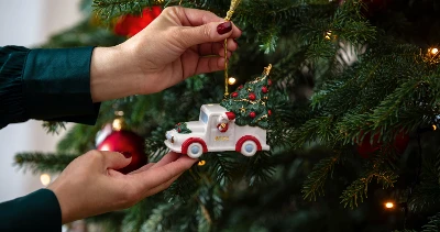 A person hangs a small truck-shaped ornament from Villeroy  and Boch on a decorated Christmas tree adorned with red baubles and lights.