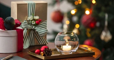 A burning candle in a Toy's Delight tea light holder A burning candle in a Toy's Delight tea light holder stands on a tray next to wrapped presents, with a decorated Christmas tree in the background.