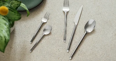 Cinq couverts like. Like. by Villeroy & Boch - fourchette, couteau, cuillère, fourchette à dessert et cuillère à café - sont disposés sur une surface en pierre claire à côté d'une assiette verte et d'une plante.	