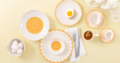 Tischgedeck mit Villeroy & Boch Fleur Soleil-Tellern, Eier, Tee mit einer Zitronenscheibe und weißen Blumen auf hellgelbem Hintergrund.