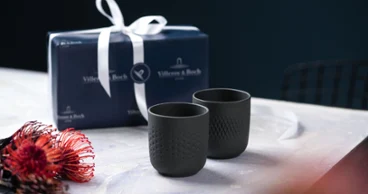 Deux tasses à collier noires sur une table avec un paquet cadeau, à côté d'une fleur rouge et noire.