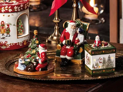 The decorative “A Christmas Toy” tray from Villeroy & Boch features ceramic Santa Claus figures, a Santa sitting on a chair, a Santa holding a tree, and a Santa sitting on a box. To the left is a Christmas-themed container.