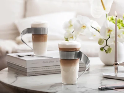 NewWave - Enjoy coffee elegantly Two layered lattes in modern NewWave glass mugs with curved metal handles stand on a marble table with books, flowers, and candles.