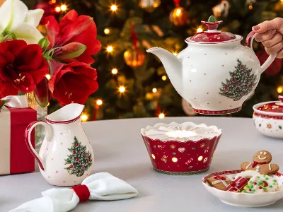 Ein festlicher Tisch mit einer Teekanne, einem Stövchen, einer Tasse, weihnachtlicher Keramik, Plätzchen und einem geschmückten Baum im Hintergrund.
