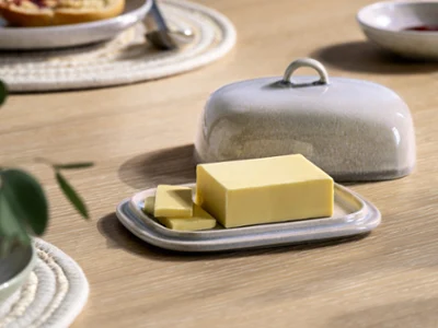 Un blocco di burro rettangolare con due fette giace su una burriera in ceramica Perlemor con coperchio abbinato su un tavolo di legno. 