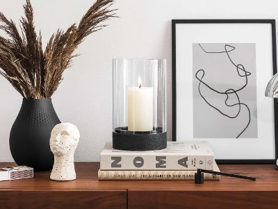 A modern highboard with decorative objects, including a vase with dried plants, a black Manufacture hurricane lamp, a sculpture, stacked books and a piece of framed abstract artwork.