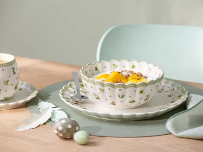 Un bol décoratif Easter Delight contenant des fruits et du yaourt est posé sur une assiette avec une cuillère, à côté d'une tasse et d'une soucoupe, sur une table.