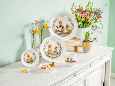Colección Spring Fantasy sobre un aparador de comedor Cuatro cuencos de Pascua con motivos de conejitos, una taza de Pascua y unas macetas con flores de primavera, todo ello sobre un aparador de madera blanca