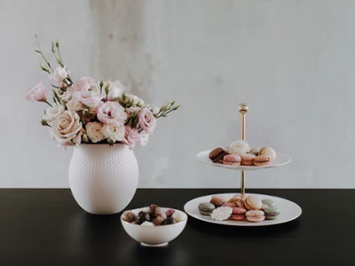 Vase Manufacture et présentoir MetroChic Un vase blanc avec des fleurs roses, un plateau à plusieurs étages avec des desserts et une coupe de chocolats sur une table sombre.