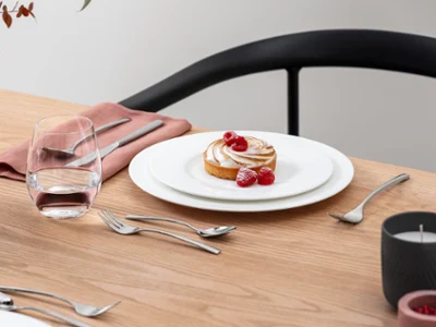 Koninklijk gecombineerd met zoete lekkernijen Een dessert gegarneerd met frambozen staat op een koninklijk bord met bestek op een houten tafel. Ernaast staat een glas water, een roze servet en een bloemstuk.