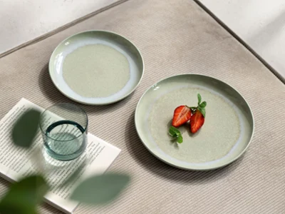 Perlemor Alga & Like Sage – Subtle Green Harmony Vista de mesa en primer plano con platos verde suave Perlemor Alga de Villeroy & Boch, decorados con un higo partido por la mitad como acento natural. El conjunto se completa con un vaso de agua Like Sage, creando un ambiente sereno y refinado donde las formas orgánicas y los tonos terrosos se unen en perfecta armonía.