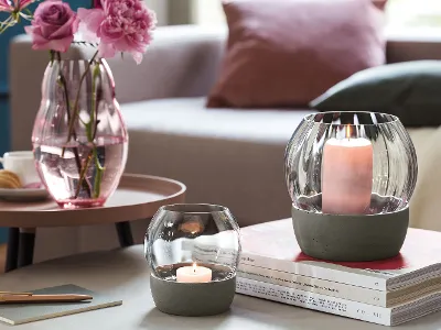 Zwei Rose Garden Windlichter von Villeroy & Boch stehen auf einem Stapel Bücher neben einer rosa Rose Garden Vase mit Blumen, alles auf einem runden Tisch in einem gemütlichen Wohnzimmerambiente.	