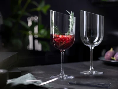 Copa NewMoon con sugerente bebida Una copa NewMoon de Villeroy & Boch decorada con líquido rojo, fruta y una ramita de romero se encuentra junto a una copa vacía sobre una mesa oscura.