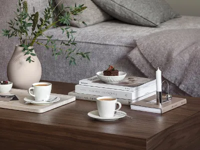 Ein gemütlicher Wohnraum mit einem hölzernen Couchtisch, auf dem zwei Manufacture Tassen Kaffee, ein kleines Dessert, ein Bücherstapel, eine Kerze und eine Manufacture Collier Vase mit Grünpflanzen stehen. 