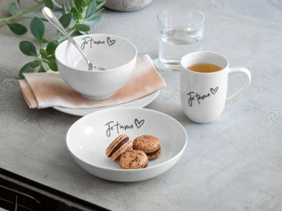 Juego de desayuno «Statement» de Villeroy & Boch Juego de desayuno «Statement» de Villeroy & Boch compuesto por un bol, un plato y una taza, cada uno con la inscripción «Je t'aime». Hay macarons en el plato y una cuchara en el bol. Un vaso de agua completa la escena, con una planta al fondo.