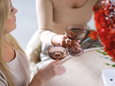 Deux personnes tiennent des verres Like Glass de Villeroy & Boch remplis de boissons et sont assises à une table décorée d'une composition florale rouge et d'un livre portant l'inscription « Mallorca ».	