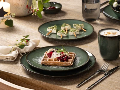 Lave est idéal pour Noël dans des tons naturels Une assiette verte Lave avec une gaufre à la crème et aux baies, une tasse de café Lave et des brochettes d'apéritifs sur une table en bois décorée pour les fêtes.