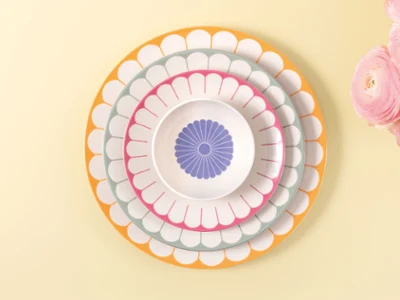 Cuatro platos Fleur de Villeroy & Boch de distintos tamaños y colores sobre un fondo amarillo claro junto a flores rosas.