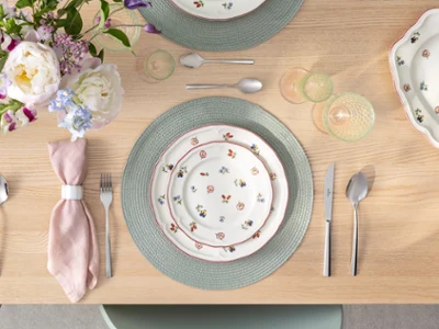Une table dressée avec des assiettes à fleurs Petite Fleur, des couverts, une serviette rose, un set de table vert et un bouquet de fleurs sur une table en bois. 