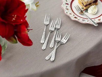 Five dessert forks on a beige tablecloth next to a plate with cake and whipped cream and a vase with red flowers.