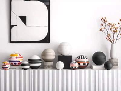 Divers La Boule de différentes tailles et couleurs Une série de La Boule sphériques, colorées, rayées et à motifs, est posée sur une console blanche sous une œuvre d'art abstrait et un vase de fleurs orange.