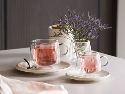 Des tasses en verre Artesano de Villeroy & Boch remplies de thé rose, placées sur des soucoupes à côté d'une cuillère sur une table décorée d'un petit vase de fleurs violettes.