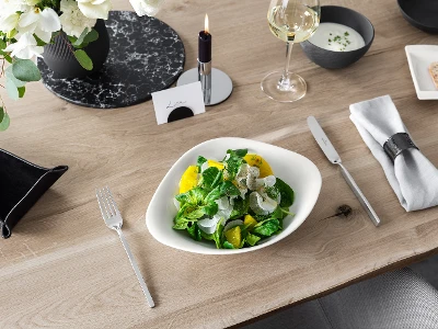 A Vapiano bowl from Villeroy & Boch with salad on a wooden table, next to it a knife and fork from NewWave.