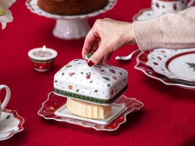 A hand lifting the lid of a festive Villeroy & Boch "Toy's Delight" butter dish on a red tablecloth, surrounded by crockery with a Christmas design.