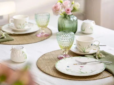 Das Set Colourful Spring von Villeroy & Boch verschönert jeden Tisch für zwei mit Tellern und Tassen mit Blumenmuster, leuchtend grünen und gelben Gläsern, rosa Blumen in einer Vase und beigen Tischsets auf einer weißen Tischdecke.