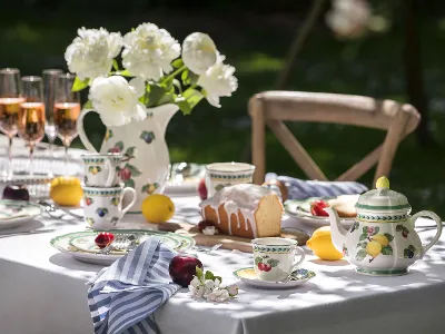 Gedekte tafel buiten met gebloemd French Garden servies, een theepot, rosé glazen en een taart. Witte pioenrozen in een vaas en fruit zijn rond de tafel geplaatst.