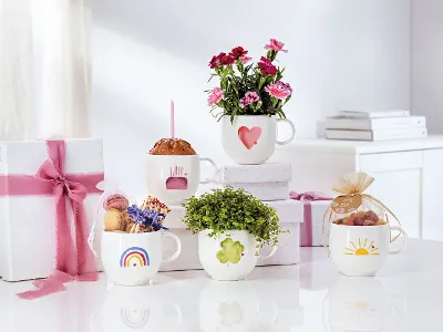 Five white “With Love” cups from Villeroy & Boch, each with a different flower, candy, and gift motif on a white surface. Two presents wrapped in pink ribbons complete the enchanting scene in the background.