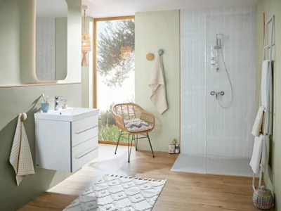 Une salle de bains moderne avec un lavabo mural, un grand miroir, une chaise en osier et une douche blanche. Des serviettes de couleurs neutres sont suspendues à des crochets et la pièce est éclairée par une grande fenêtre qui diffuse une lumière naturelle.