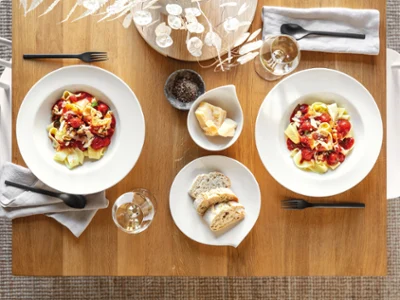 Des assiettes à pâtes Flow sur une table pour 2 personnes Deux assiettes Flow de Villeroy & Boch contenant des pâtes, des tomates et du fromage sur une table en bois, accompagnées d’un plat creux contenant du pain, de deux verres à vin, de couverts de table et d’un centre de table décoratif.