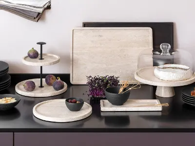 Küchenarbeitsplatte mit eleganten Manfacture Travertin Tabletts, auf denen Feigen, Brie, Brot und eine Schüssel Gemüse präsentiert werden. 