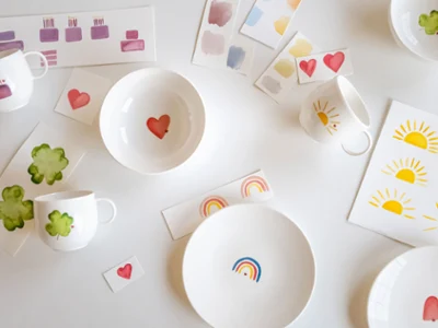 Juego de platos y tazas blancos "With Love" de Villeroy & Boch, decorados con motivos alegres Un juego de platos y tazas blancos "With Love" de Villeroy & Boch, decorados con animados corazones, arco iris y motivos arbóreos, elegantemente colocados junto a patrones de colores y tarjetas de patrones sobre una superficie blanca inmaculada.