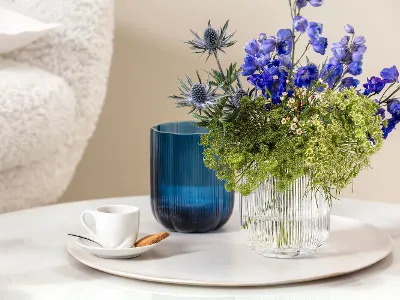 A small white For me cup with a biscuit on a saucer, next to a blue Fleur vase and a transparent Fleur vase with blue and green flowers on a round marble surface.