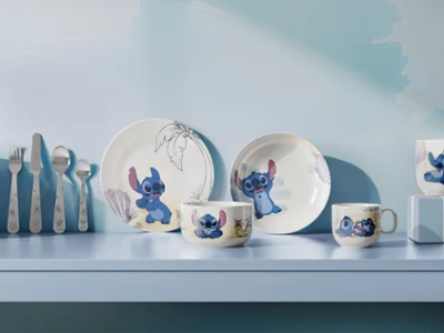Una serie di piatti e tazze della collezione Disney Stitch sono decorati con motivi dei cartoni animati ed esposti su uno scaffale azzurro insieme ad altri utensili.