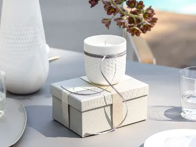 Une bougie blanche Manufacture Blanc avec un ruban gris repose sur une boîte cadeau fermée sur une table. À proximité, deux verres d'eau et un vase Manufacture Blanc.