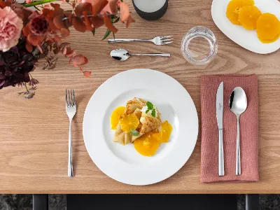 An elegantly set dining table with a plate of crêpes topped with orange slices and mint. Cutlery is placed next to a glass and a bowl with more orange slices.