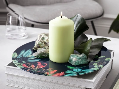 A still life scene with a lit candle, crystals and foliage on a decorative Avarua plate, with books and a pen on a modern table.