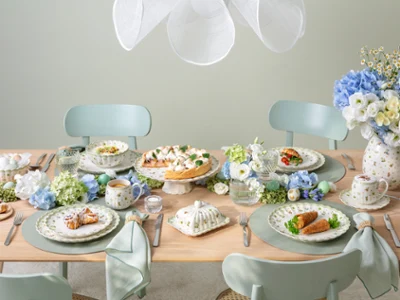 Une table à manger pour quatre personnes décorée dans des tons pastel, avec des assiettes remplies de plats, des tasses à thé, des compositions florales bleues et blanches et une vaisselle élégante, créant une atmosphère fraîche et accueillante.