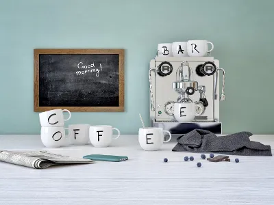 Espresso machine with Villeroy & Boch "Letters" cups spelling out the word "COFFEE" on a table, next to a phone, a newspaper, blueberries and a blackboard with the words "Good morning!".