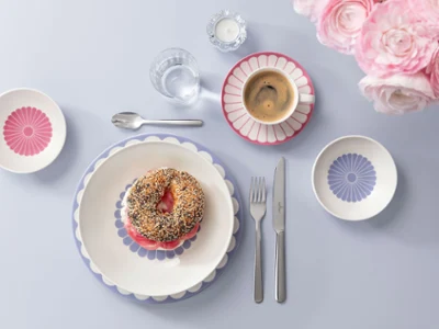 Service de table avec un bagel sur une assiette décorée. Service de table avec un bagel sur une assiette décorée, entouré d'une tasse à café, de petits bols et de couverts sur une surface bleu clair avec des fleurs roses sur le côté.