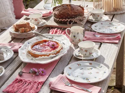 The Villeroy & Boch Petite Fleur collection is elegantly arranged on a wooden table and includes floral-patterned plates, a cake, a tart, cups and pink napkins for tea.