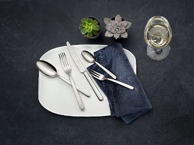Cubiertos Louis en plato blanco con servilleta azul Servicio de mesa con un plato blanco, cubiertos de acero inoxidable de la colección Louis, una servilleta oscura, dos pequeñas suculentas y una copa de vino blanco sobre una superficie oscura.
