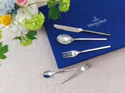 Besteckset mit Messer, Löffel und Gabel auf blauem Tuch neben Blumenarrangement auf beigem Untergrund.	