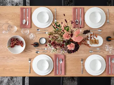 Montaje de mesa floral y uvas Una mesa de comedor de madera con platos blancos, cuencos, cubiertos sobre servilletas rosas, dos velas, un bol con uvas y un arreglo floral.