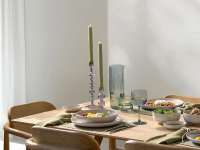 A wooden dining table with plates full of food, glasses, green candles and minimalist crockery.