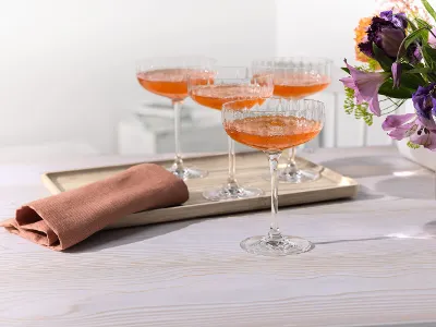 Four glasses filled with orange cocktails on a tray stand next to a folded napkin and flowers on a light-colored table.