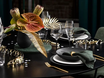 Une élégante présentation de table de Villeroy & Boch avec des assiettes Manufacture foncées, des couverts de table dorés, des serviettes vertes et un vase Collier noir avec des fleurs et des feuilles tropicales. Des serpentins et des verres dorés décorent la table noire.